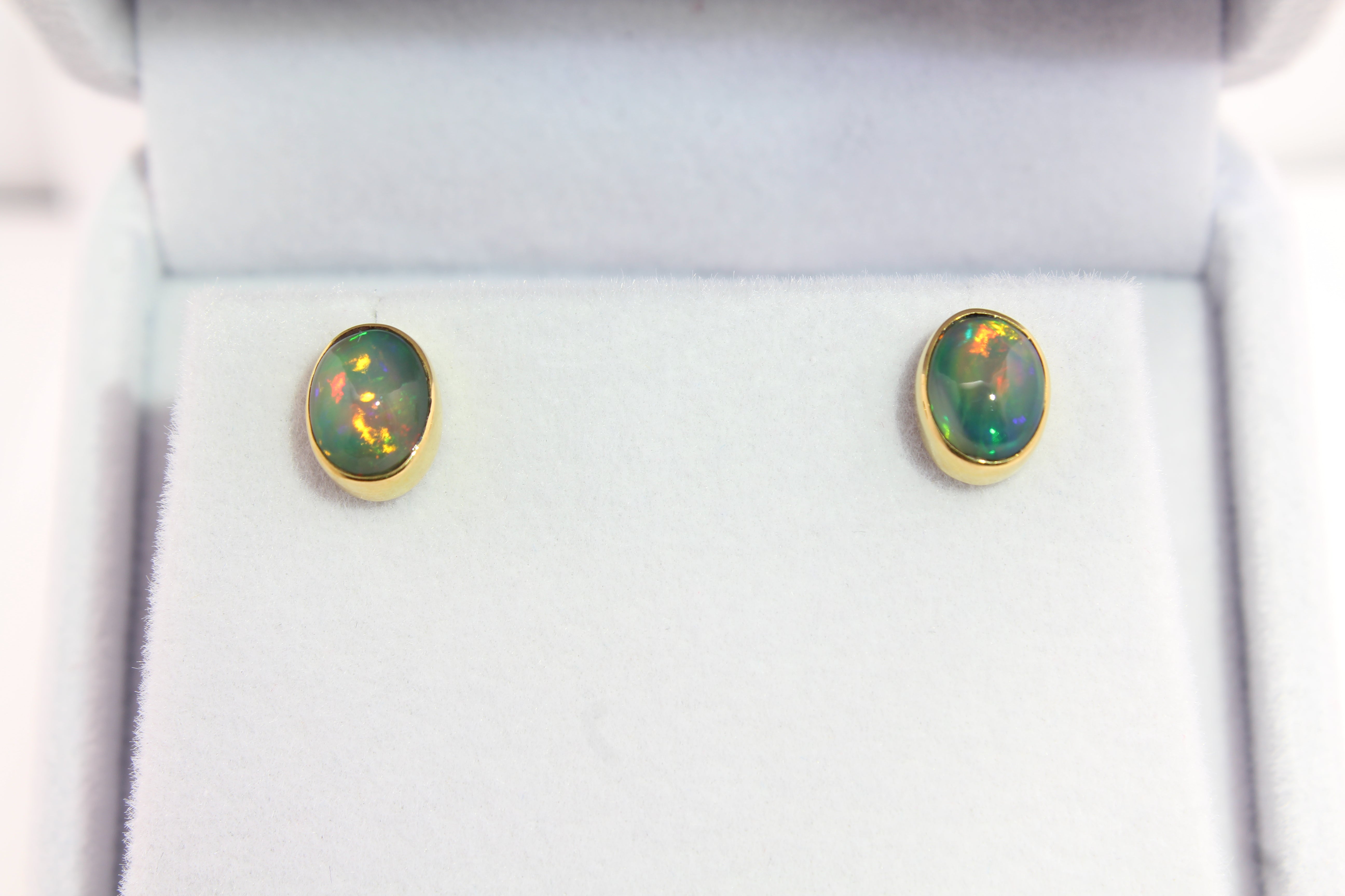 Pair of oval Ethiopian opal stud earrings featuring vibrant flashes of orange, red, and gold, set in handcrafted 22k gold bezels on a white display.
