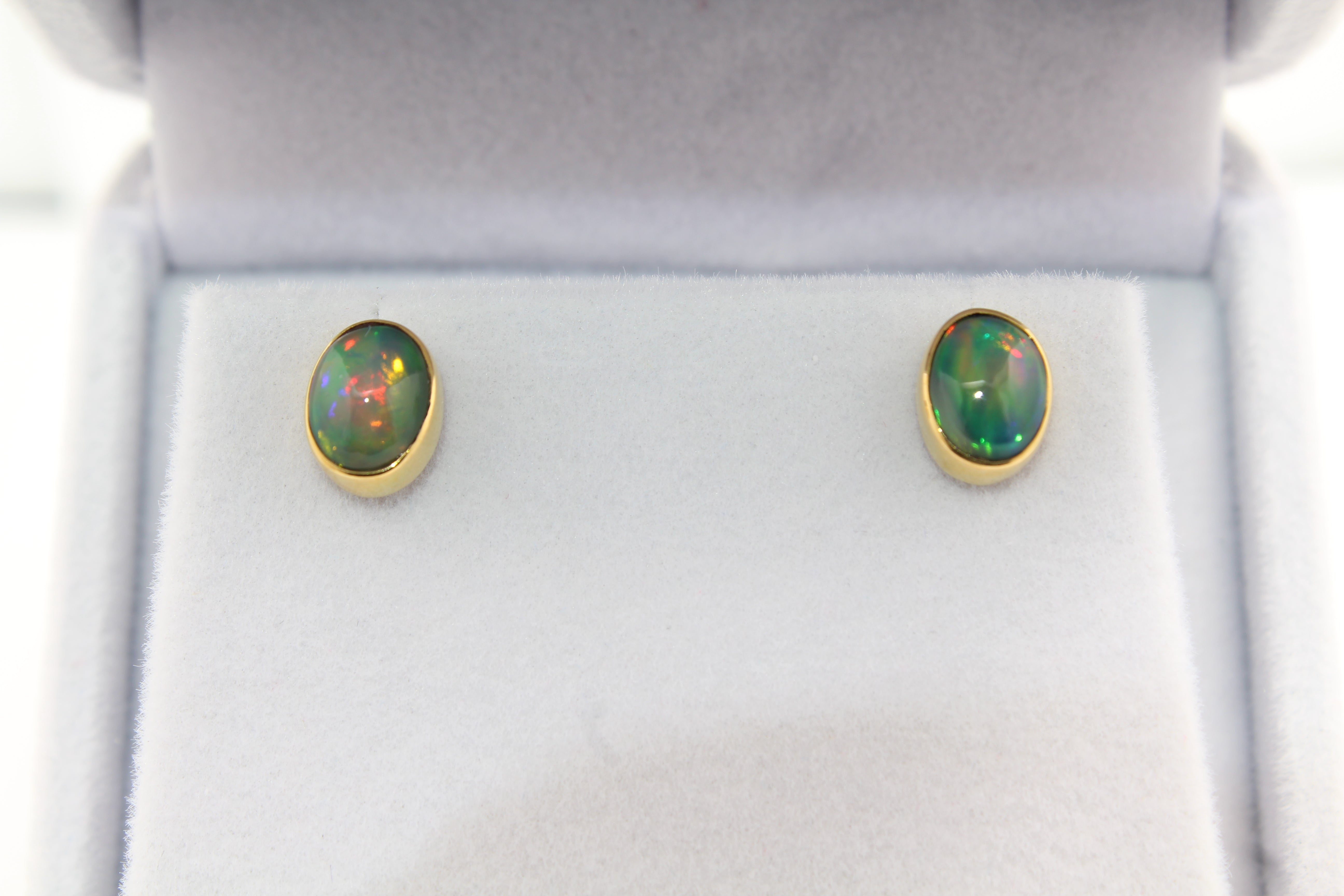 Pair of oval Ethiopian opal stud earrings featuring vibrant flashes of green, purple, and red, set in handcrafted 22k gold bezels on a white display.
